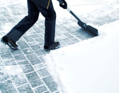 A person shovels snow off a paved walkway, leaving a clear path. They are wearing dark pants and boots.