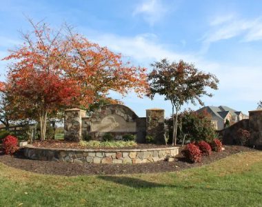 Stone entrance sign surrounded by autumn trees and shrubs, under a clear blue sky, with houses visible in the background.