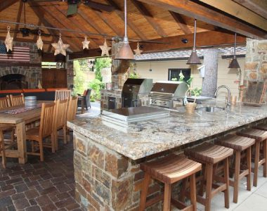 Outdoor kitchen with stone countertops, bar stools, and a wood-fired oven. Dining table with chairs under a wooden pergola. Grills and sink in the background. Hanging lights and decorations.