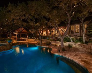 Night view of a backyard with a lit swimming pool, surrounded by trees and a stone patio. A house with large windows and outdoor seating is visible in the background.
