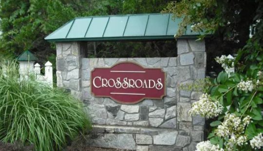 Stone sign with "Crossroads" in white text on a red background, surrounded by greenery.