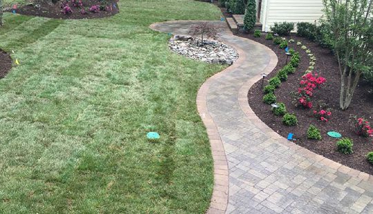 A curved stone path in a garden with neatly trimmed grass, a small rock-lined area, and flower beds along the path.